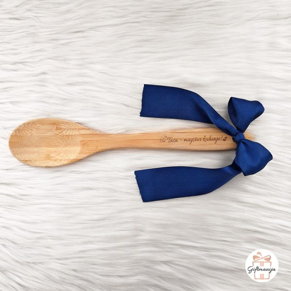 Drvena kuhača s plavom mašnom i ugraviranim natpisom Tata majstor kuhinje, položen na bijeloj čupavoj podlozi.