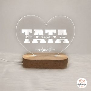 Akrilna 3D lampa za tatu s imenima djece urezanim unutar slova riječi TATA, u obliku srca na drvenom postolju, svijetli toplim svjetlom.