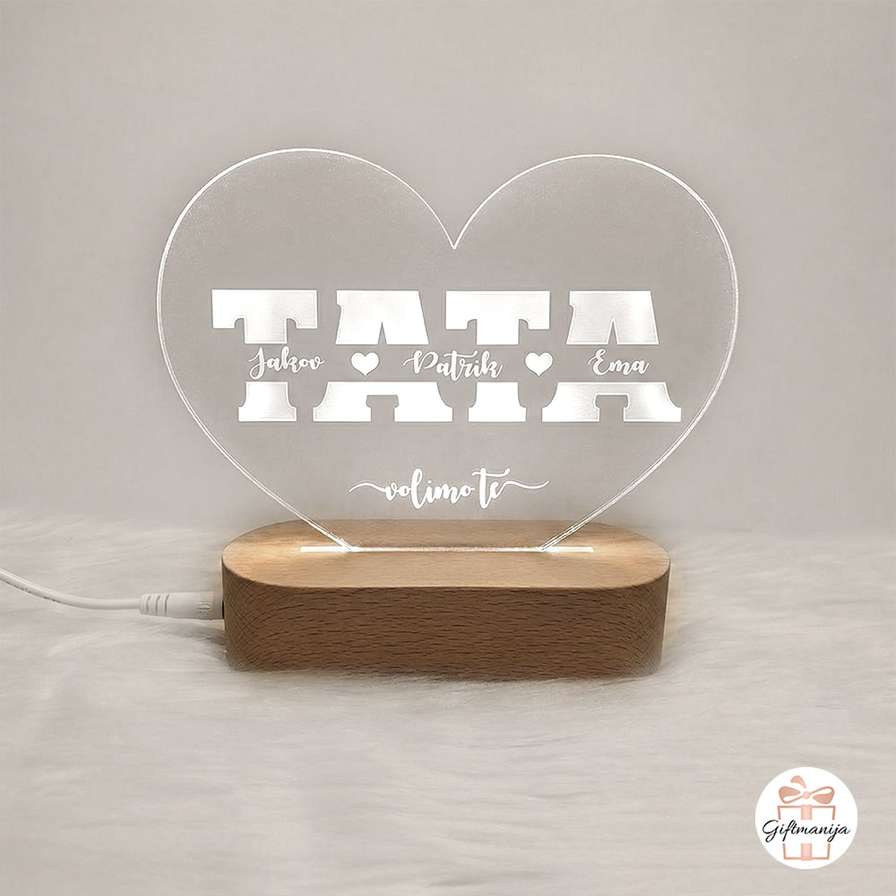 Akrilna 3D lampa za tatu s imenima djece urezanim unutar slova riječi TATA, u obliku srca na drvenom postolju, svijetli toplim svjetlom.